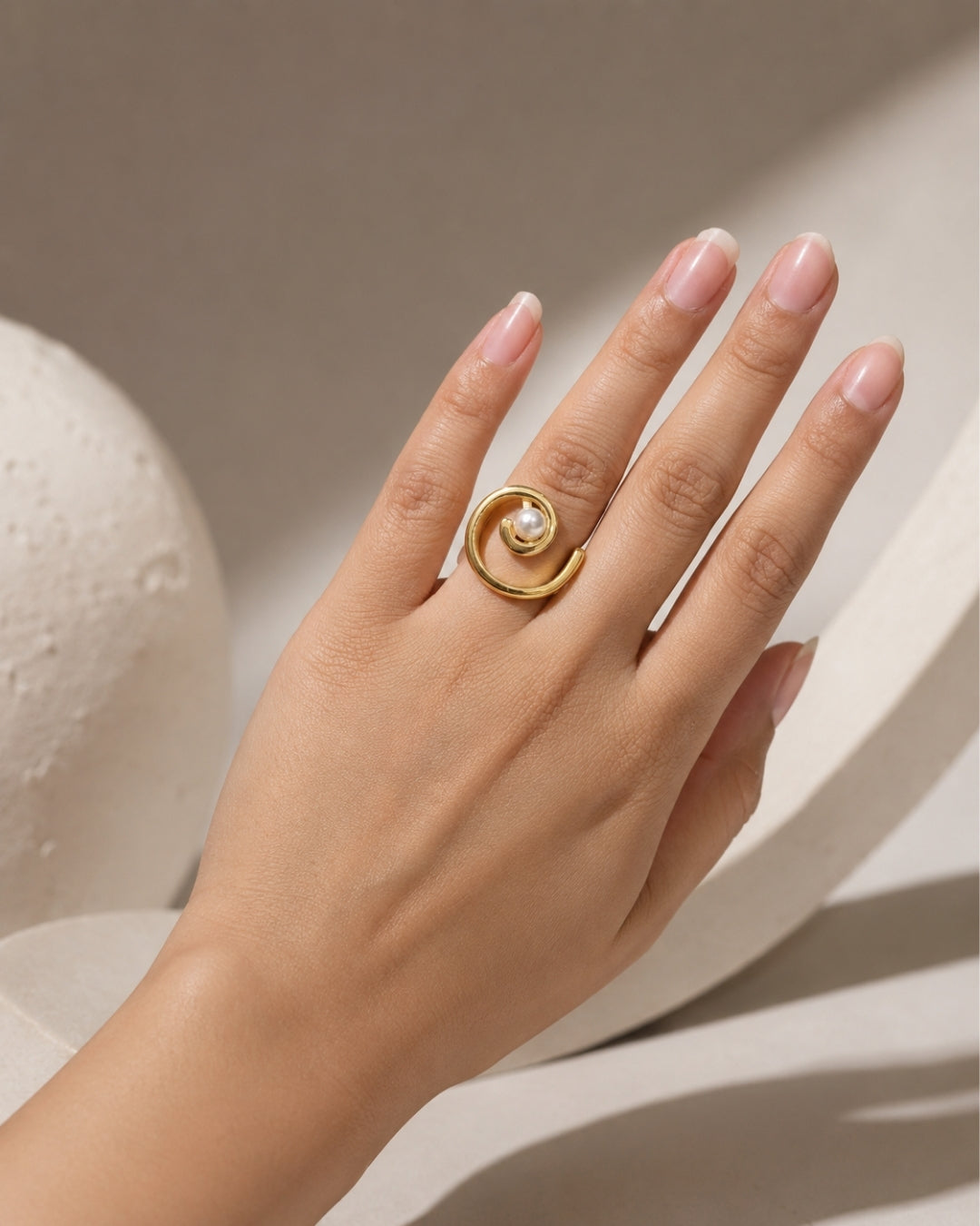 A gold spiral ring with a central white pearl, worn on a hand with manicured nails, set against a minimalist beige background with soft shadows.