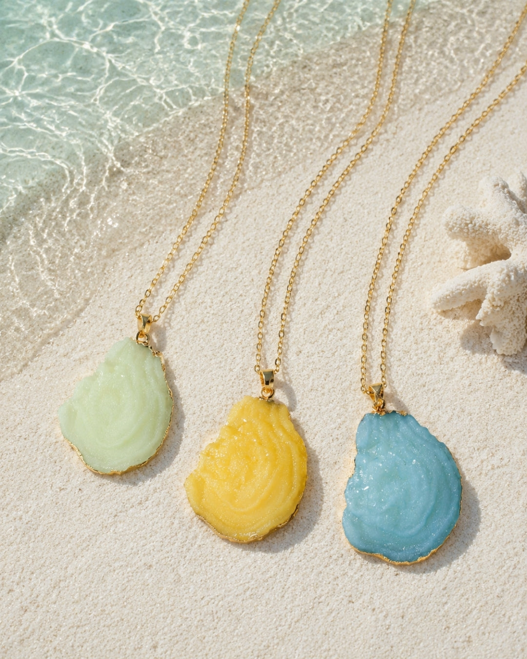 Three gold chain necklaces with irregular polished stone pendants in pale green, yellow, and light blue, arranged on a sandy beach with shallow water and a starfish.