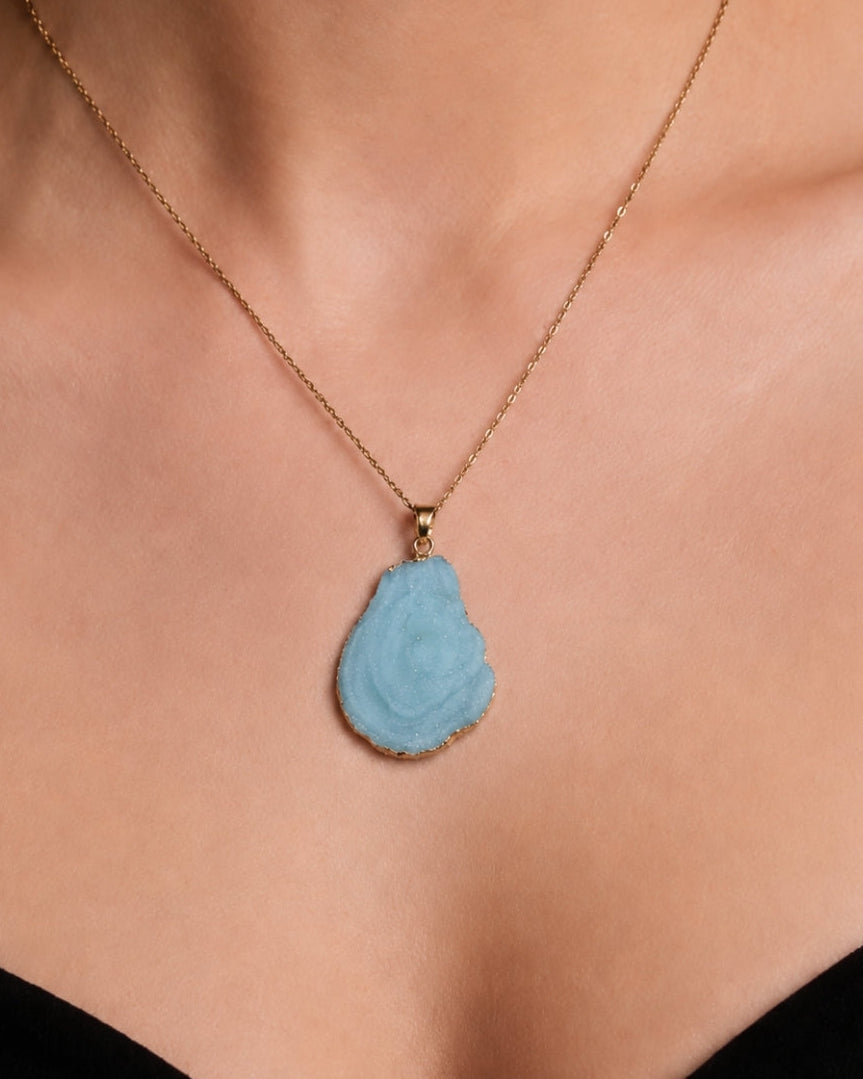A close-up of a model's neck and chest wearing a delicate gold chain necklace with a carved, teardrop-shaped light blue pendant.