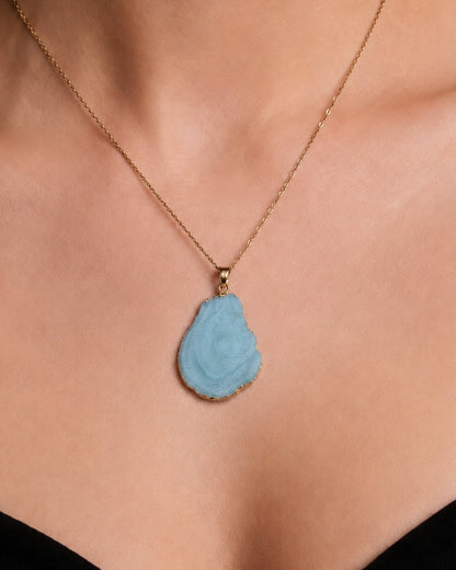 A close-up of a model's neck and chest wearing a delicate gold chain necklace with a carved, teardrop-shaped light blue pendant.