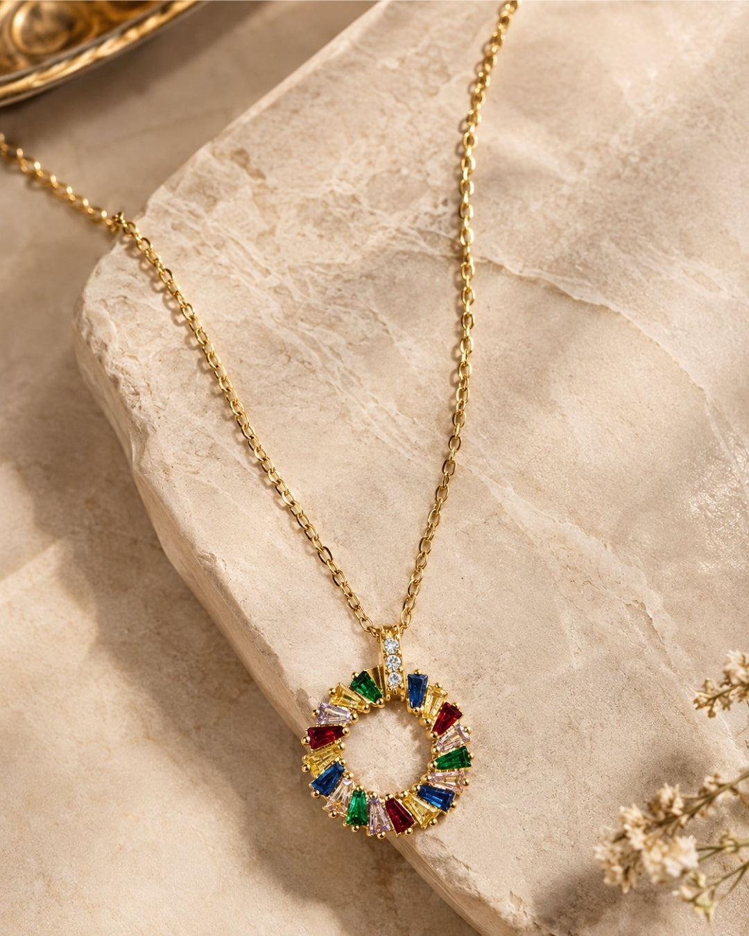 A gold chain necklace with a circular pendant set with colorful gemstones, displayed on a textured beige stone slab.