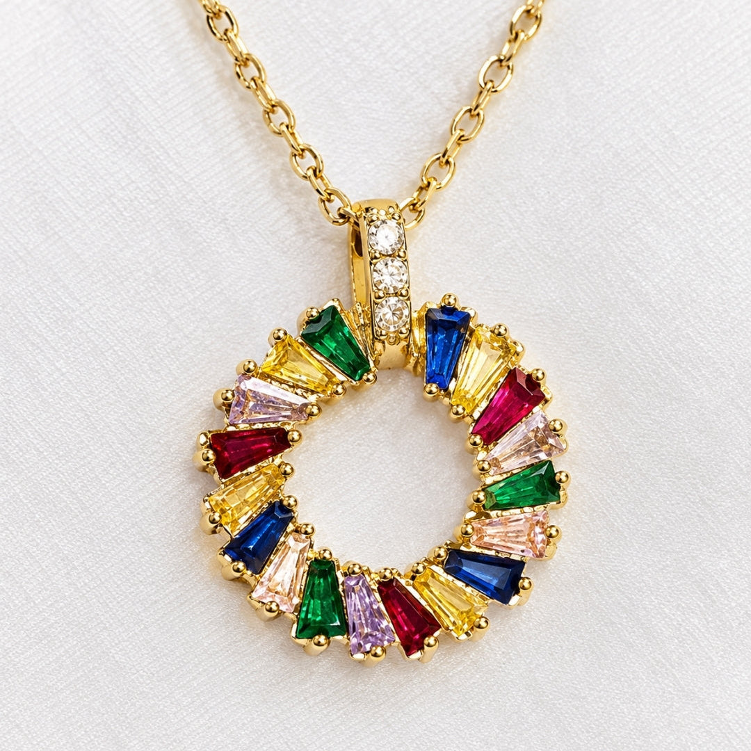 A gold chain necklace with a circular pendant set with colorful baguette gemstones in yellow, blue, green, pink, and red, displayed on a textured white fabric background.