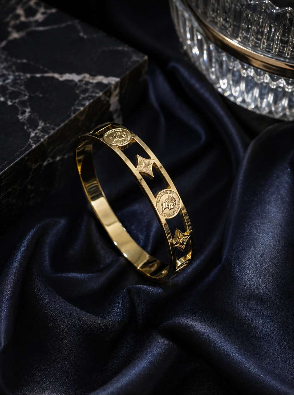 A polished gold bangle bracelet with engraved circular medallions and star motifs, displayed on draped dark navy satin fabric next to a black marble block.