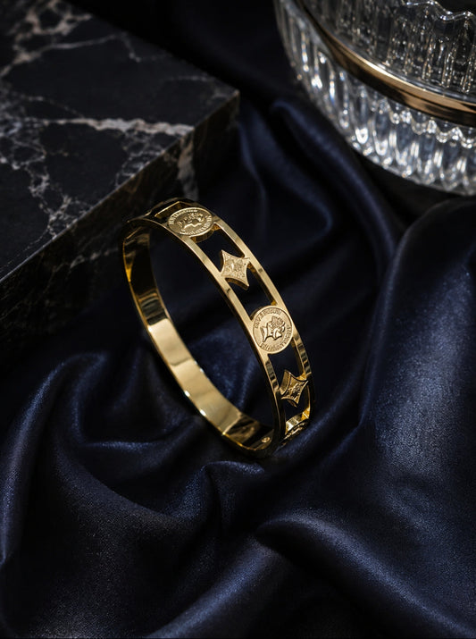A polished gold bangle bracelet with engraved circular medallions and star motifs, displayed on draped dark navy satin fabric next to a black marble block.