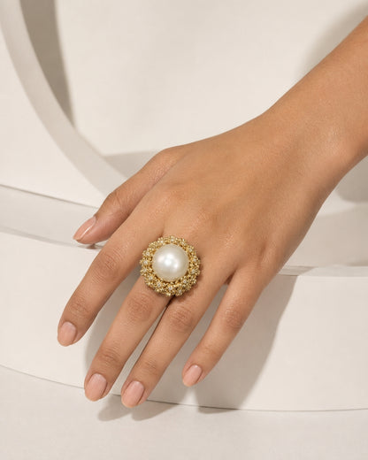 A close-up of a hand wearing a large statement ring with a white pearl surrounded by a halo of small gold and clear stones, set against a clean white architectural background.