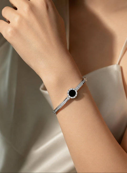 A close-up of a model's wrist wearing a silver-toned bangle bracelet with a central round black stone surrounded by small clear stones, paired with a satin cream dress.