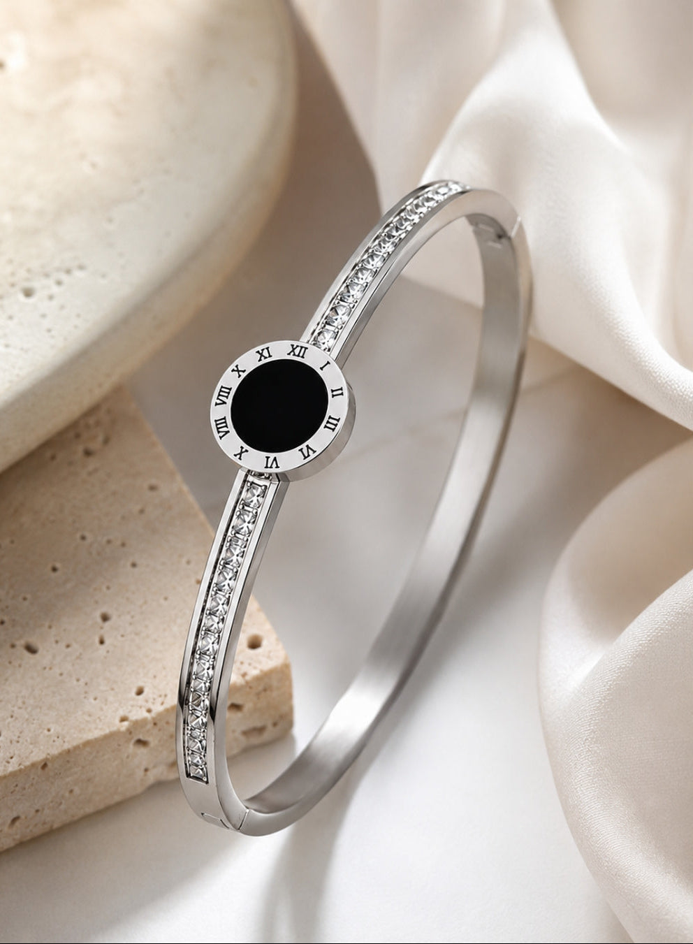 A polished silver-toned bangle bracelet with a central round black inlay and Roman numeral markers, accented with small clear stones along the band, styled on a white satin cloth with a beige stone prop.