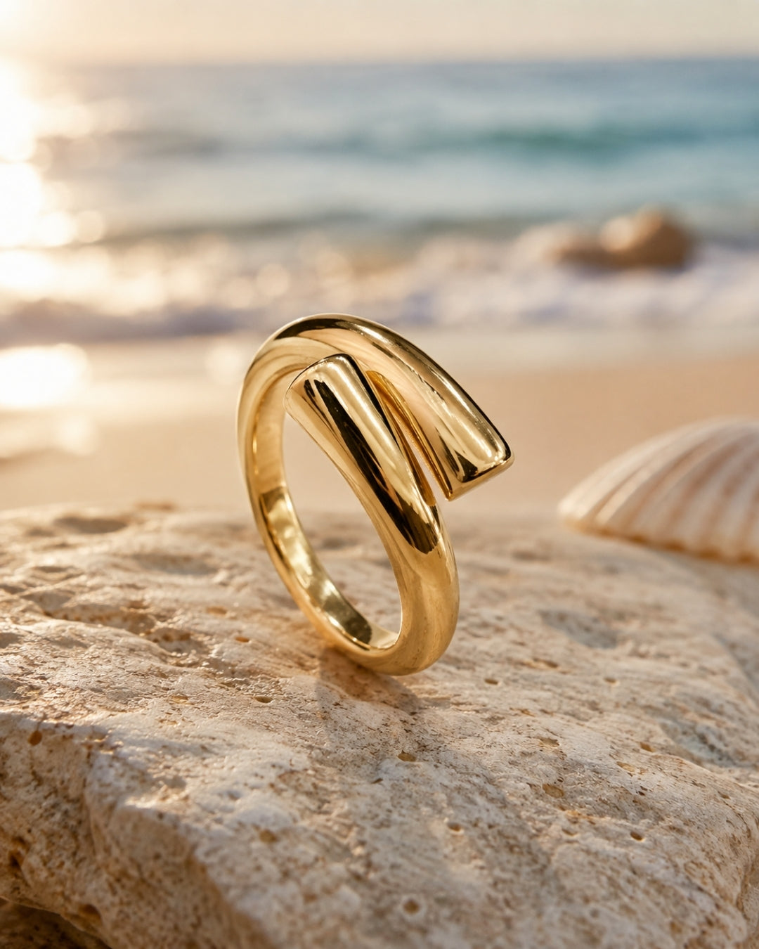 A polished gold ring with a bold, sculptural wrap design, resting on a textured beach stone with a seashell and the ocean in the background.