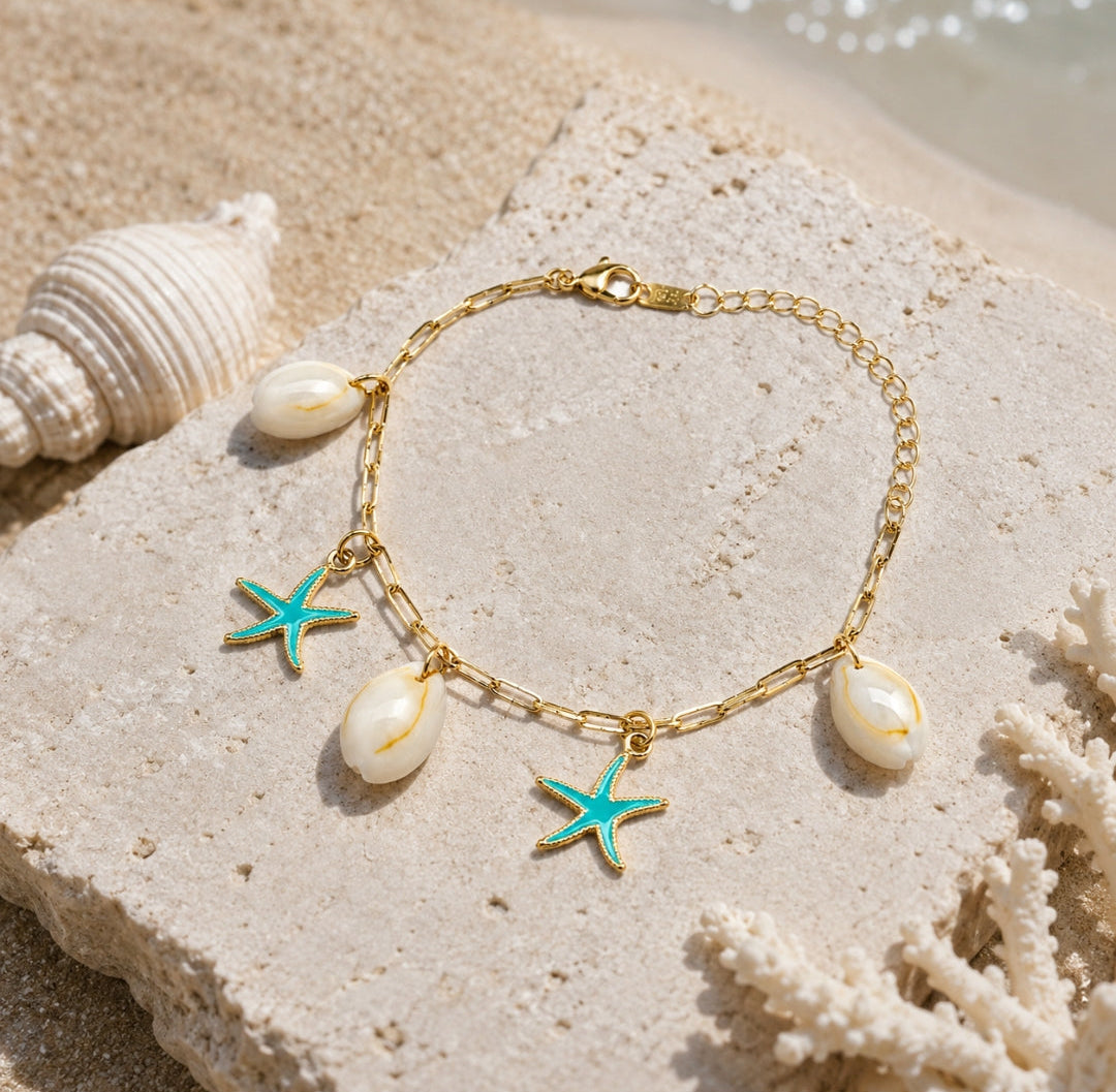 A gold chain bracelet with three white cowrie shells and two turquoise starfish charms, displayed on a textured stone with seashells and sand in the background.