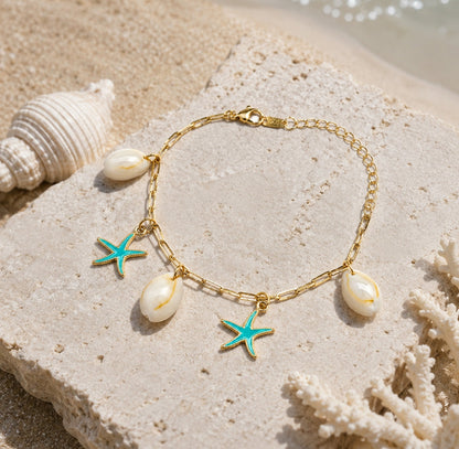 A gold chain bracelet with three white cowrie shells and two turquoise starfish charms, displayed on a textured stone with seashells and sand in the background.