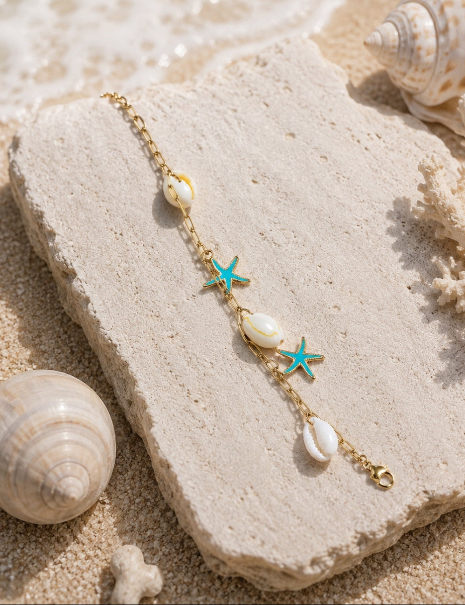 A gold chain bracelet with white seashells and turquoise starfish charms, displayed on a textured stone block over a sandy beach setting with real shells and coral.