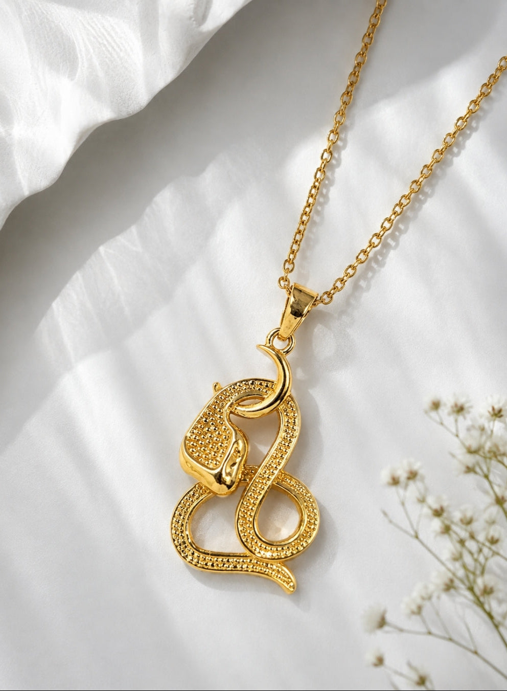 A gold pendant necklace featuring a coiled snake design with textured pavé-style detailing, displayed on a soft white draped fabric background with small white flowers.
