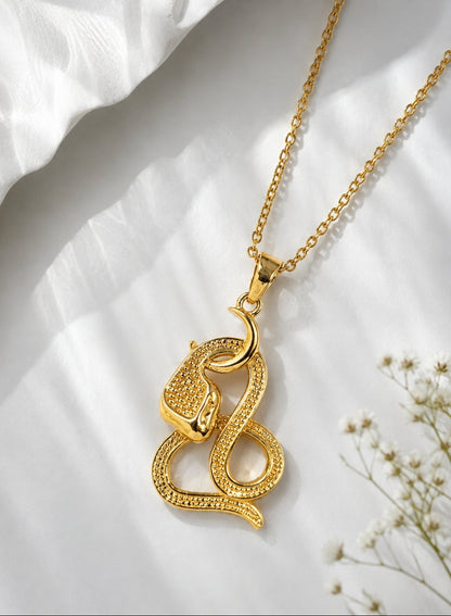 A gold pendant necklace featuring a coiled snake design with textured pavé-style detailing, displayed on a soft white draped fabric background with small white flowers.
