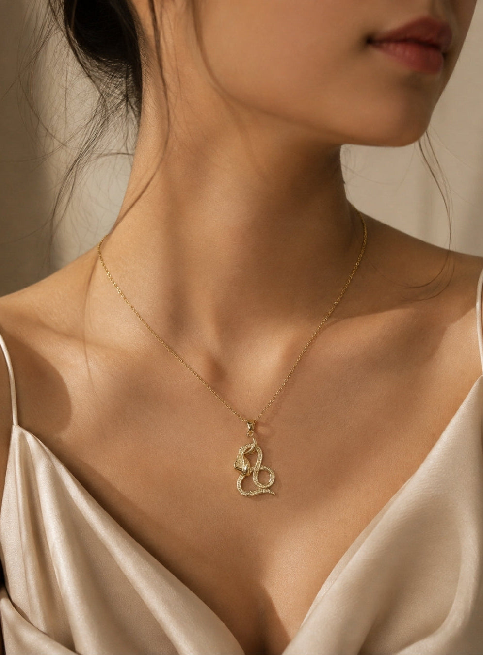 A close-up of a woman wearing a delicate gold chain necklace with a coiled snake pendant, styled with a satin cream top.