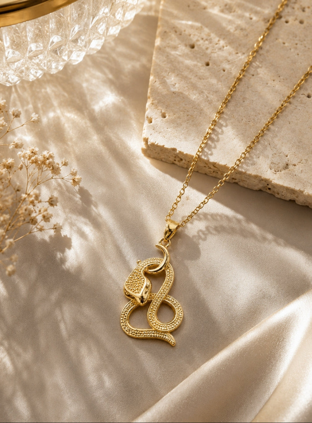 A gold snake pendant necklace with a textured, beaded snake body, resting on draped champagne-colored satin fabric next to a stone slab and dried flowers.