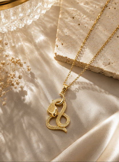 A gold snake pendant necklace with a textured, beaded snake body, resting on draped champagne-colored satin fabric next to a stone slab and dried flowers.