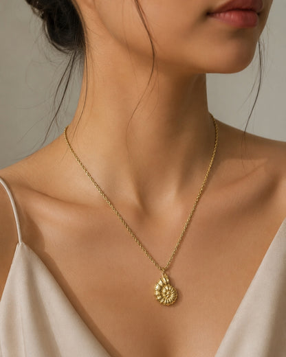 A close-up of a woman wearing a delicate gold chain necklace with a textured, spiral shell-like pendant, styled with a cream-colored top against a soft neutral background.