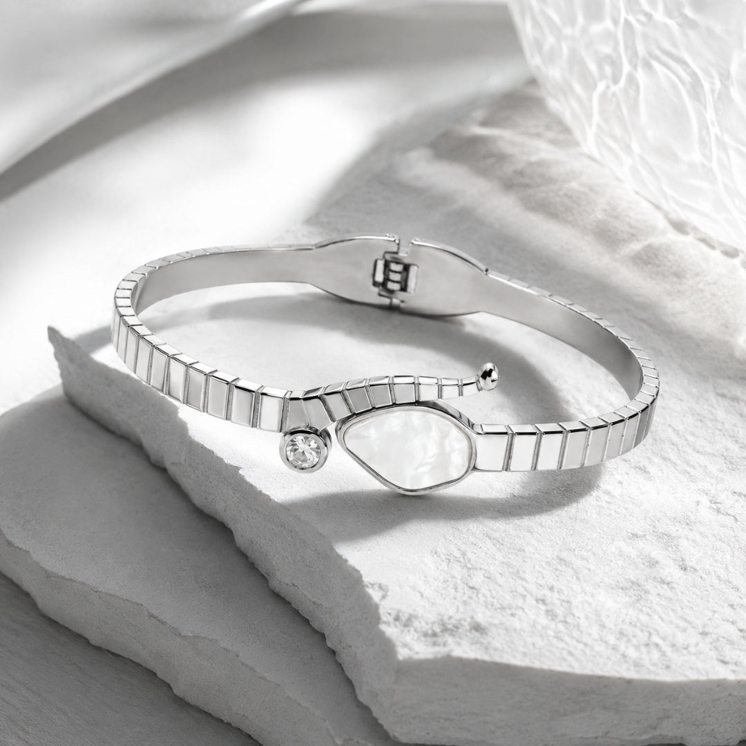 A silver-toned bangle bracelet with a textured segmented design, a smooth teardrop-shaped inlay, and a small clear stone accent, displayed on a white stone slab.