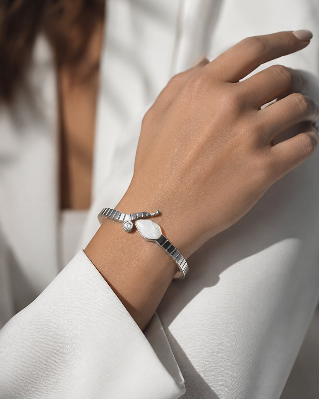 A close-up of a person's hand and wrist wearing a silver wrap bracelet with a white oval stone, styled with a white blazer.