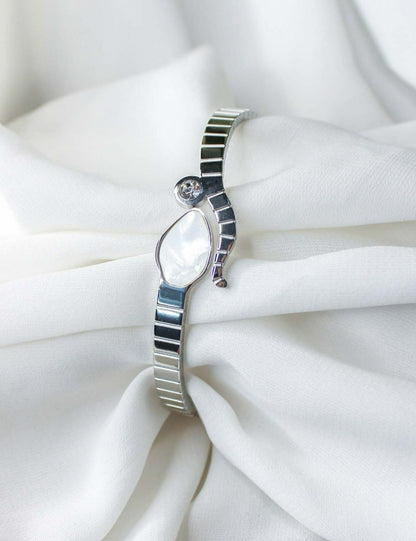 A sleek stainless steel snake bracelet with a mother of pearl embellished snake head on an open bangle, displayed on a white fabric surface.