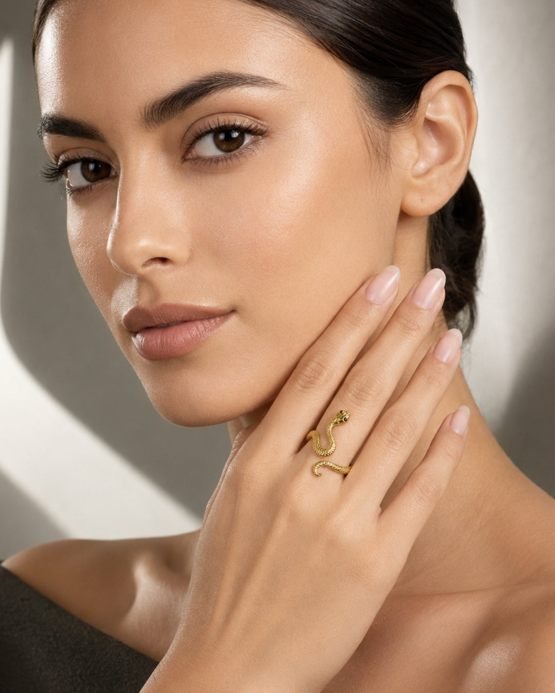 A close-up lifestyle shot of a woman with dark hair tied back, wearing a delicate gold snake ring with a small green gemstone on her ring finger.