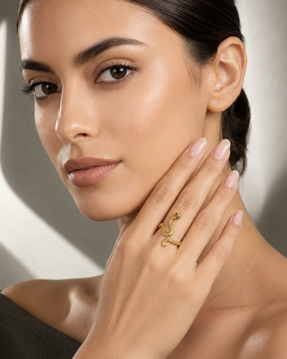 A close-up lifestyle shot of a woman with dark hair tied back, wearing a delicate gold snake ring with a small green gemstone on her ring finger.