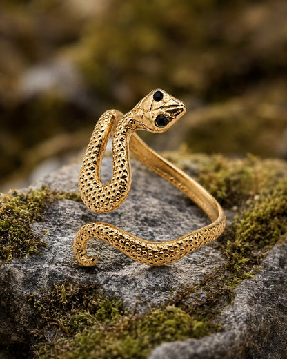 A gold snake-shaped ring with textured scales and black eyes, resting on a mossy gray rock with a blurred natural background.