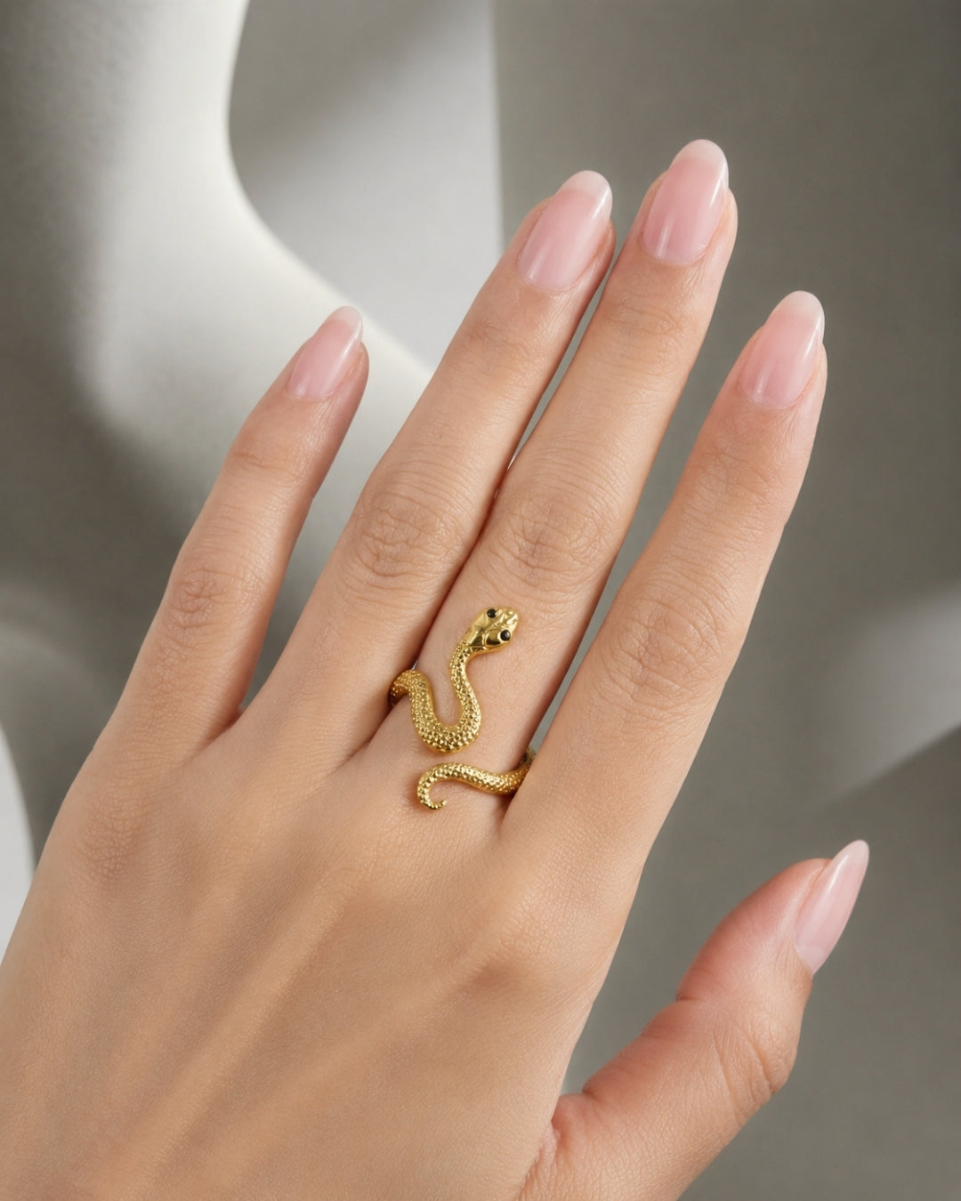 A close-up of a hand with long, neatly manicured pinkish nails wearing a small gold snake ring on the ring finger, set against a soft gray background.
