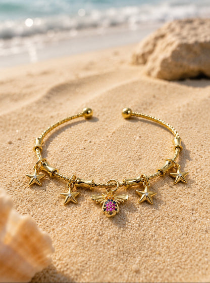 A gold-toned beaded bracelet with purple oval beads, clear spacer beads, and a circular turquoise and gold charm, styled on white sand with seashells and a blue water background.