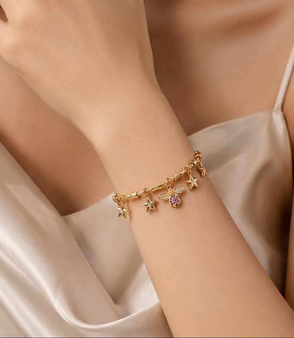 A gold-tone beaded anklet with purple and white beads, a central gold medallion featuring a turquoise and white mermaid design, and a lobster clasp, styled on white sand with seashells and a blue water background.