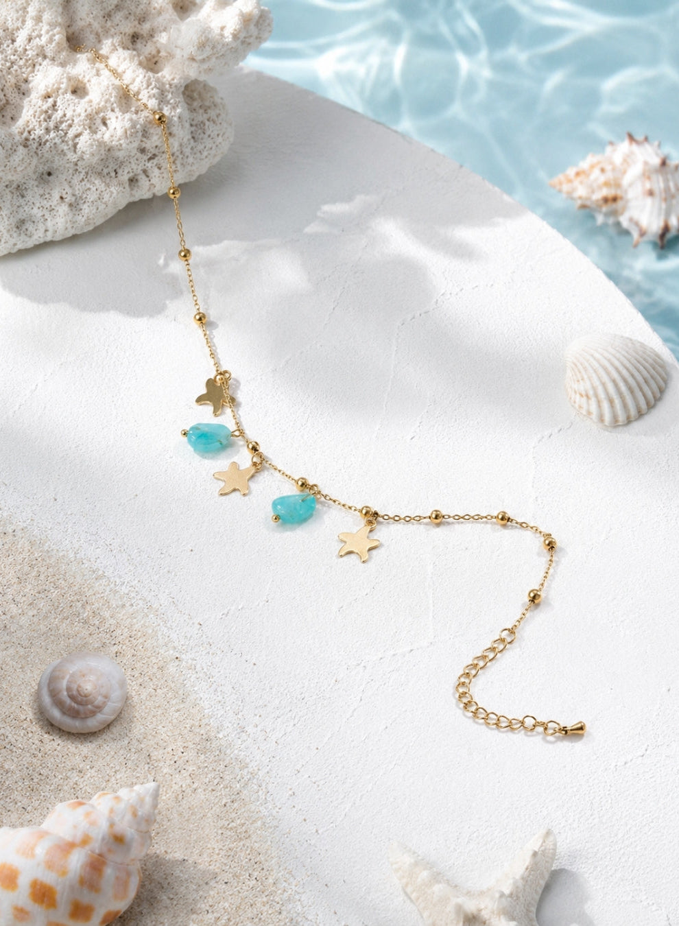 A gold-toned anklet with starfish and turquoise bead charms, styled on a white surface with seashells and a blue water backdrop.