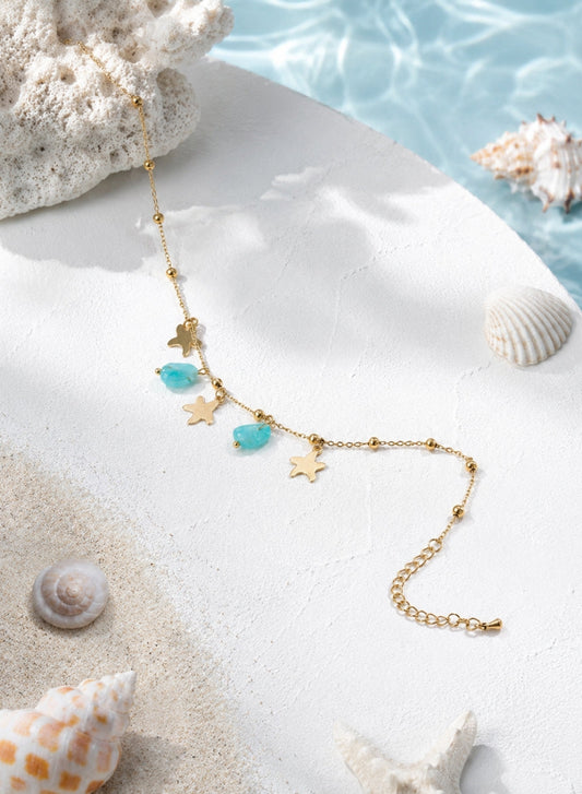 A gold-toned anklet with starfish and turquoise bead charms, styled on a white surface with seashells and a blue water backdrop.
