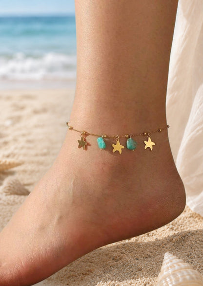 A delicate gold anklet with star and turquoise bead charms worn on a person's ankle, shown on a sandy beach with the ocean in the background.
