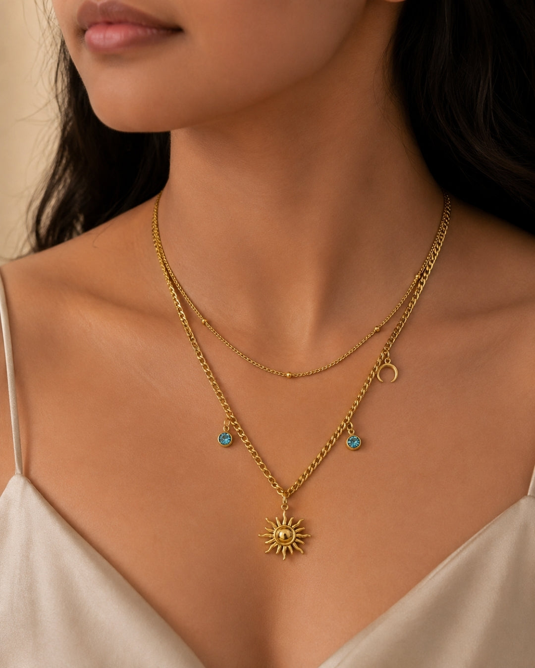 A close-up of a model wearing a double-layered gold necklace. The top layer is a delicate chain with a small crescent moon charm, while the bottom layer features a larger sun pendant and small turquoise bead charms.