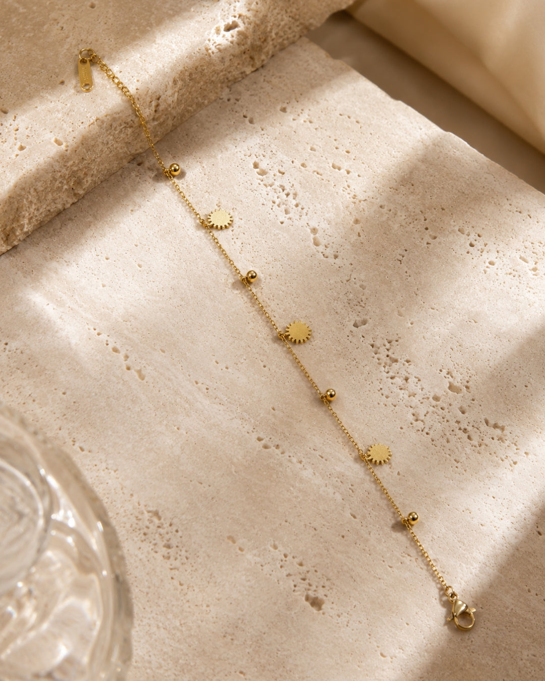 A delicate gold chain anklet with small sunburst and bead charms, laid diagonally on a textured beige stone surface with warm natural shadows.