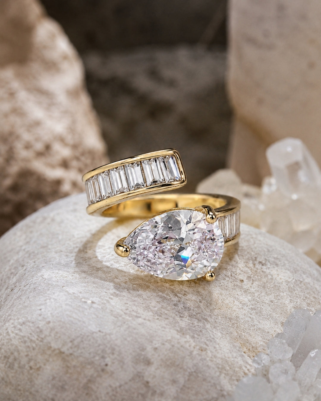 Two yellow gold rings displayed on a textured stone surface. The top ring features a channel-set row of baguette-cut clear stones, while the bottom ring has a large pear-cut clear center stone with baguette side stones.