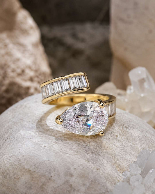 Two yellow gold rings displayed on a textured stone surface. The top ring features a channel-set row of baguette-cut clear stones, while the bottom ring has a large pear-cut clear center stone with baguette side stones.