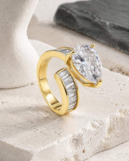 A gold ring with a large marquise-cut clear gemstone and baguette-cut side stones, displayed on a textured stone surface.