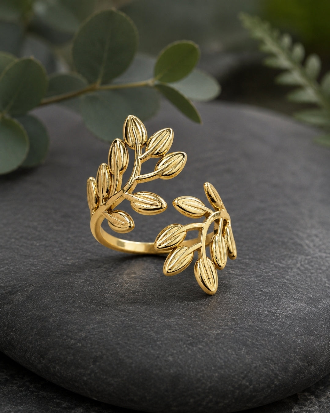 A gold leaf branch wrap ring displayed on a dark slate stone with blurred green leaves in the background.