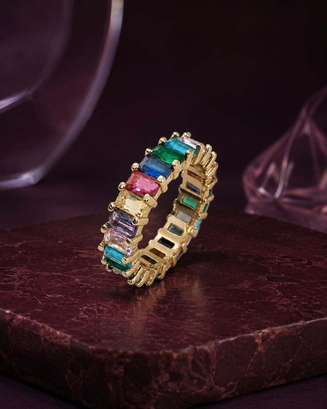 A gold eternity ring featuring a continuous row of colorful baguette-cut gemstones in green, blue, pink, purple, and clear, displayed on a dark textured stone surface.