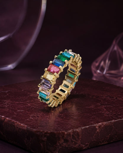 A gold eternity ring featuring a continuous row of colorful baguette-cut gemstones in green, blue, pink, purple, and clear, displayed on a dark textured stone surface.