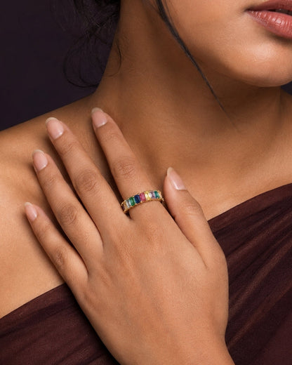 A close-up lifestyle shot of a model wearing a gold band ring with a row of colorful rectangular gemstones (green, blue, pink, orange) on her finger, set against a dark background.