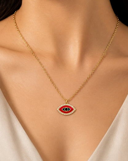 A gold chain necklace with an evil eye pendant worn by a model. The pendant features a red center, a black pupil, and a border of small clear stones.