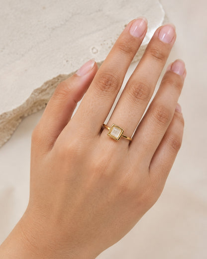 A gold ring with a square milky white stone, worn on a hand with pale pink manicured nails, set against a neutral beige textured background.