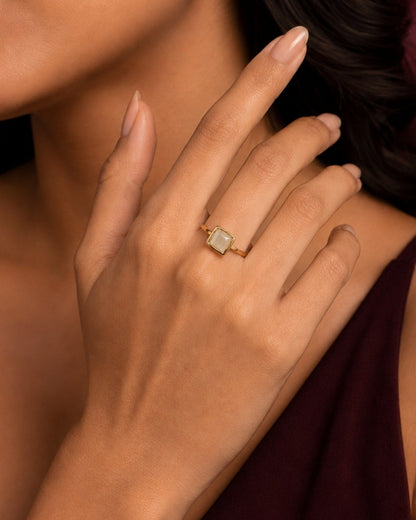 A close-up lifestyle shot of a model wearing a gold ring with a square-cut light green stone on her finger, set against a dark burgundy top and a dark background.