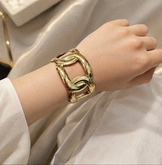 A close-up of a person's wrist wearing a wide, chunky gold-toned cuff bracelet with an interlocking open-link design, set against a soft beige satin fabric background.