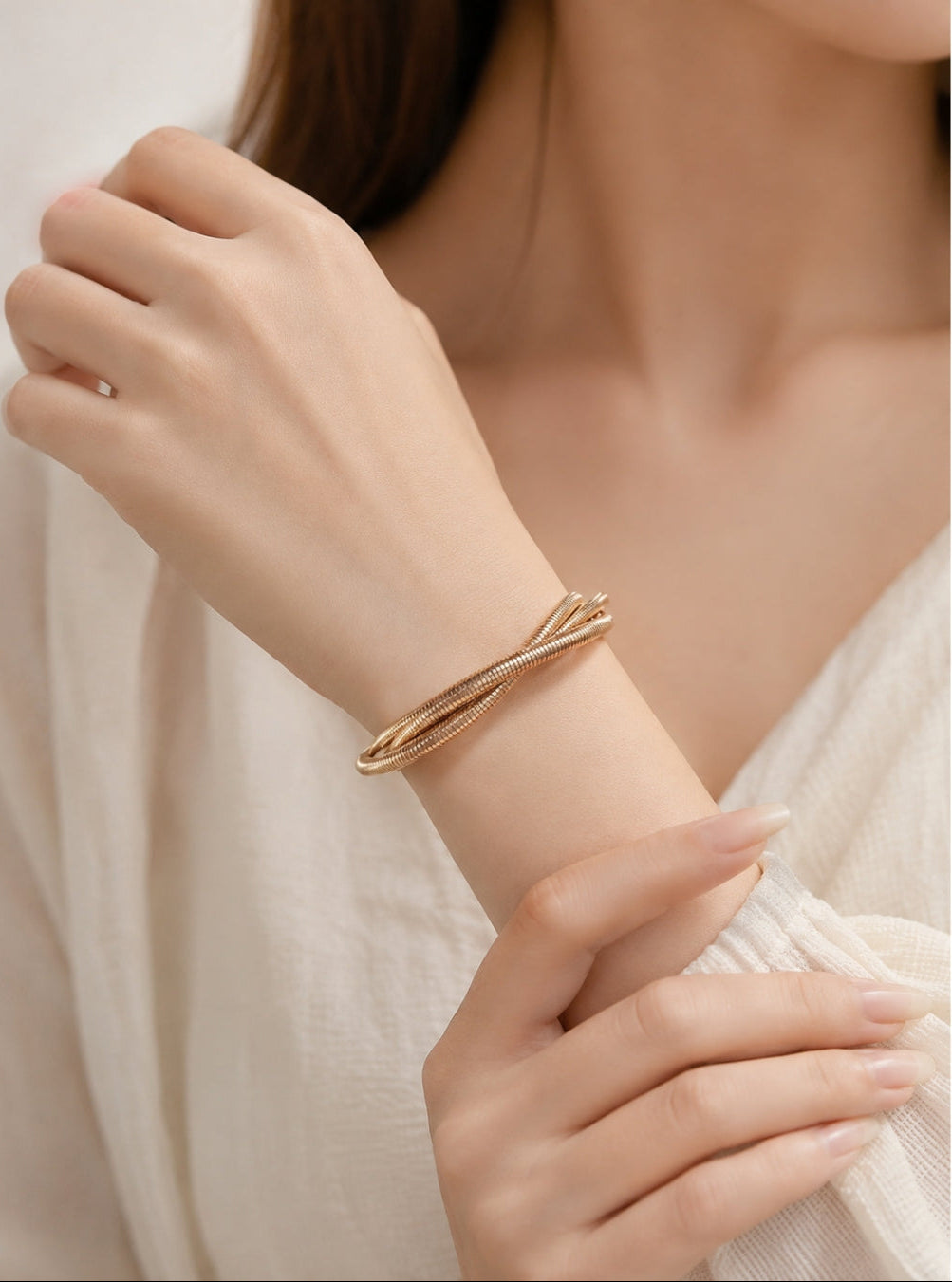 A close-up of a woman's wrist wearing a slim, twisted gold bangle bracelet, paired with a cream-colored textured top.