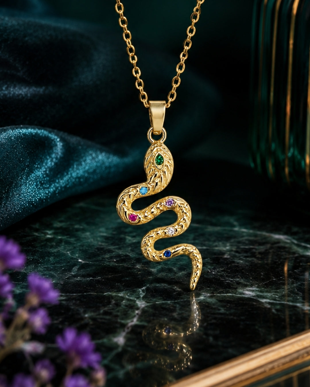 A gold snake pendant necklace with small multicolored gemstones, displayed on a dark marble surface with a teal satin cloth and purple flowers in the background.