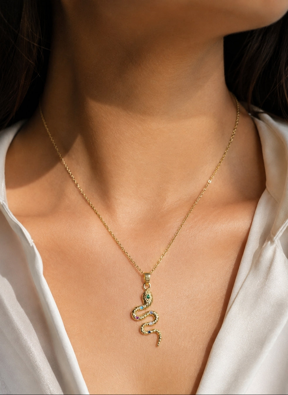 A gold snake pendant necklace with small green and pink stones, worn on a model's neck over a white blouse.