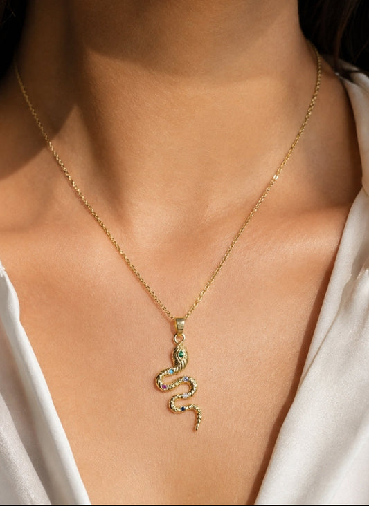 A gold snake pendant necklace with small green and pink stones, worn on a model's neck over a white blouse.
